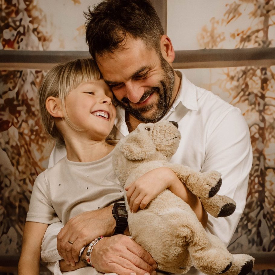 Papa und Tochter  lächeln, umarmt ein Stofftier vor einer winterlichen Kulisse. Familienfotografie Berlin.