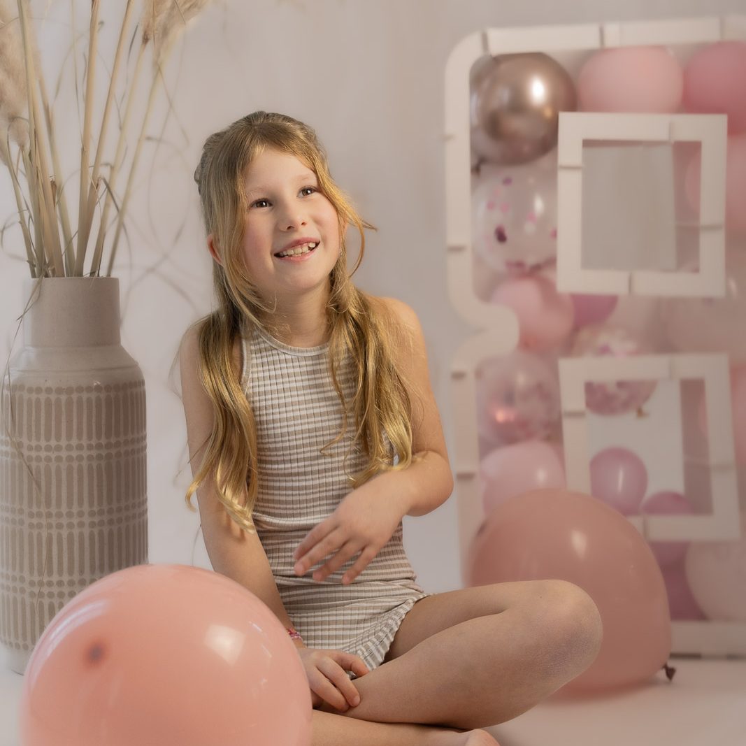Ein lächelndes Mädchen sitzt mit Luftballons in Zartrosa und einem dekorativen Hintergrund.